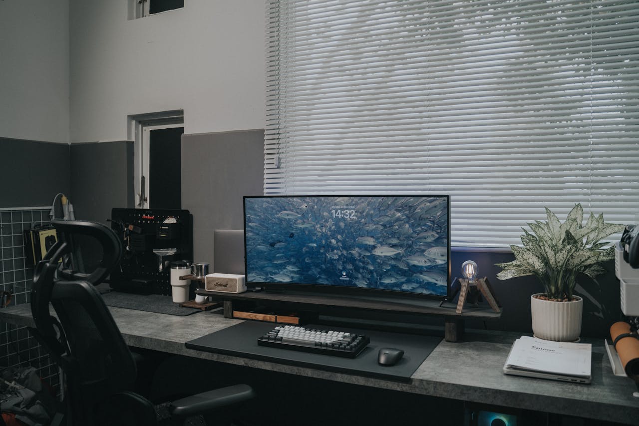 Stylish office desk featuring a large monitor, keyboard, and plant decor against a window with blinds.